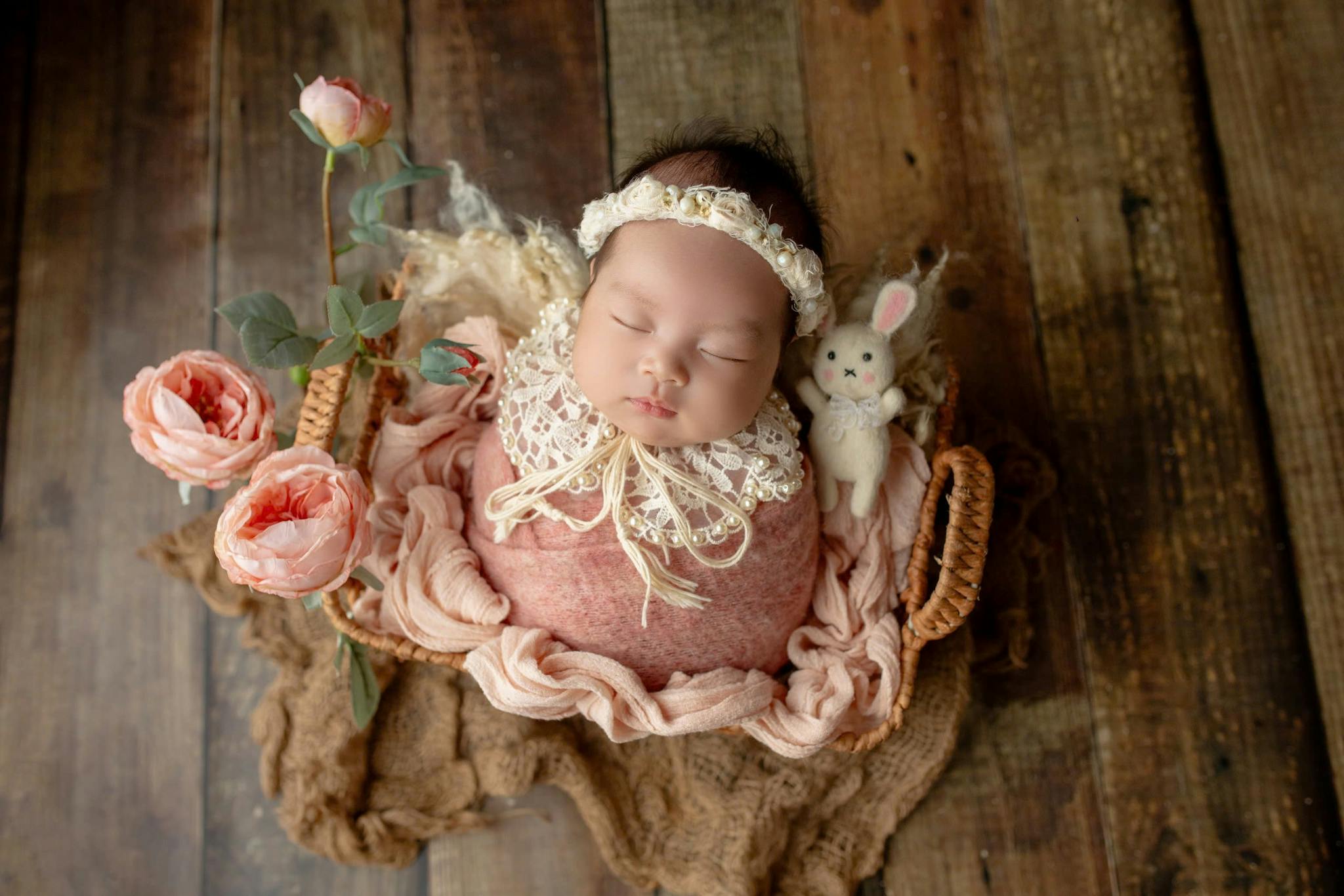 Home 6 A serene newborn baby wrapped in pink, surrounded by roses and a plush rabbit toy.