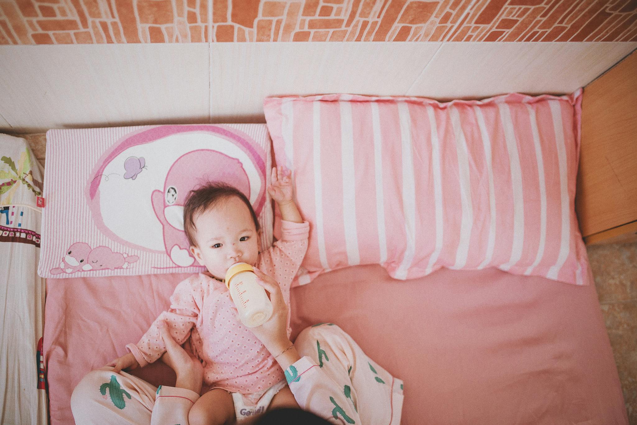 Baby lying in a pink-themed bedroom being fed with a milk bottle. Warm and soft ambiance.