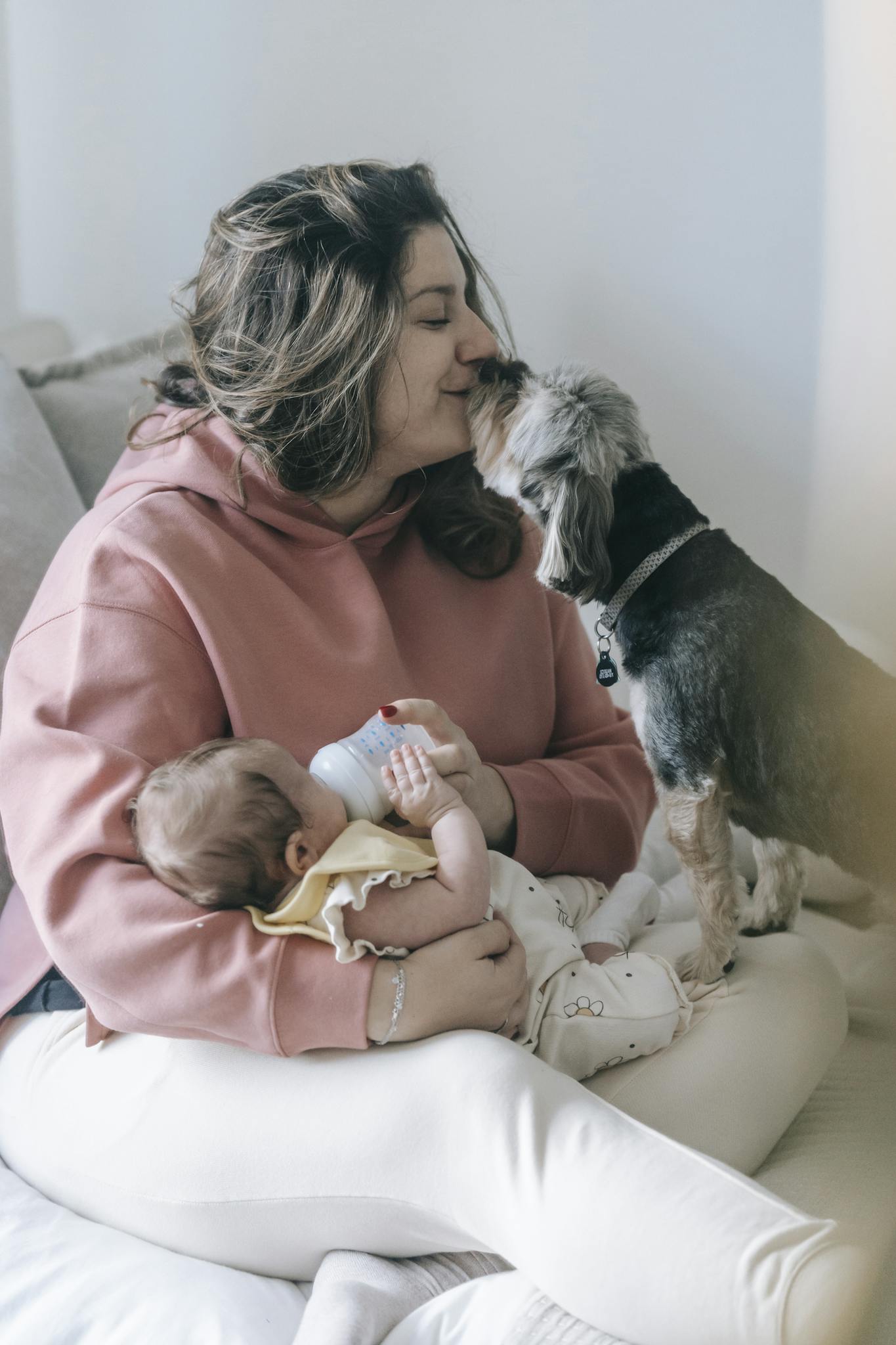 Side view of mother caressing adorable hairy dog while feeding infant baby with bottle on bed in light room at home